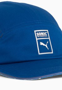Blå kasket lavet af stof, der har en firkantet lap med teksten "SONIC THE HEDGEHOG" og et Puma-logo i hvid. Flad skygge design.