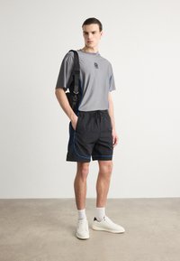 Camisa gris de manga corta con detalles en azul, combinada con pantalones cortos negros con ribetes azules. El modelo lleva deportivas blancas y calcetines blancos.