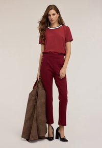 Top rosso a maniche corte con rifiniture bianche e nere, abbinato a pantaloni slim fit rossi con bottoni laterali. Il modello tiene in mano un blazer a quadri marrone.