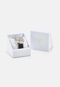 Goldfarbene Uhr mit silbernem Musterzifferblatt, schwarzem Silikonarmband, präsentiert in einer weißen strukturierten Box mit goldenen Logostickern.