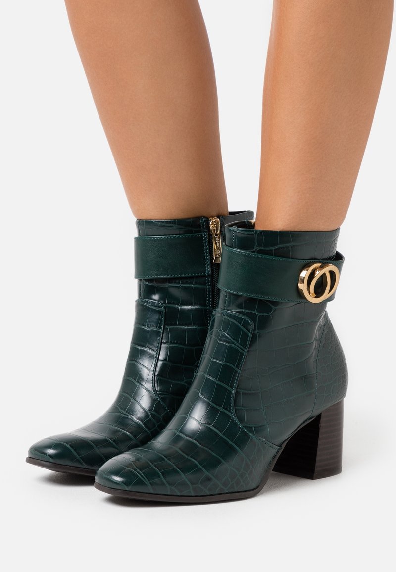 Bottes chevilles vert foncé à motif crocodile avec talons carrés, fermetures éclair latérales et détail de boucle double anneau dorée portées sur jambes nues.