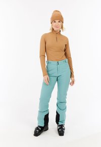 Bruine lange mouwen top met een ritskraag, gecombineerd met turquoise skibroek met een zwarte onderkant en geïntegreerde riem.
