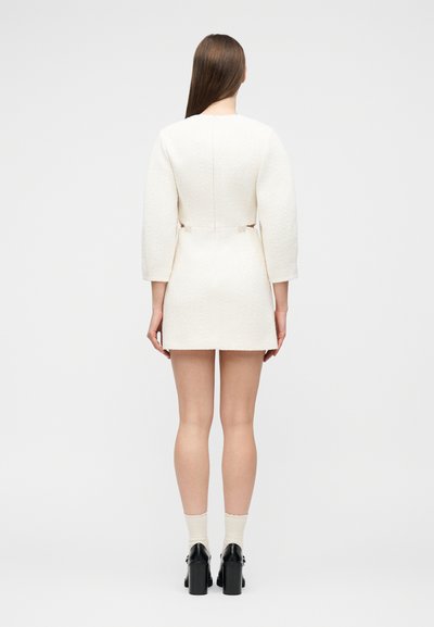 Γυναίκα με μακριά καστανά μαλλιά, ντυμένη με λευκό υφασμάτινο μίνι φόρεμα με μακριά μανίκια, κρεμ κάλτσες και μαύρα παπούτσια με ψηλά τακούνια, γυρισμένη με την πλάτη.