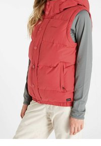 Veste matelassée rouge avec capuche, fermetures à boutons, poche latérale et un logo. Portée par-dessus un t-shirt à manches longues gris, associée à un pantalon clair.