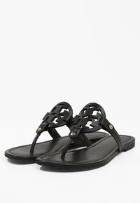 Sandales plates en cuir noir avec des lanières rondes découpées de manière complexe et de petites clous en métal doré sur un fond blanc.