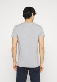 Mann mit kurzen, lockigen, dunklen Haaren, der ein schlichtes, hellgraues T-Shirt mit kurzen Ärmeln und dunkelblaue Hose trägt, und mit dem Rücken zu einer weißen Hintergrund steht.