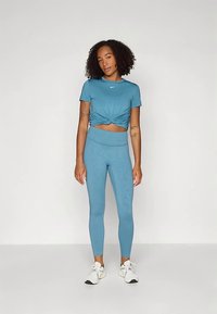 Mujer con cabello rizado que lleva una camiseta corta anudada en color verde azulado, leggings de cintura alta a juego y zapatillas blancas, de pie contra un fondo liso.