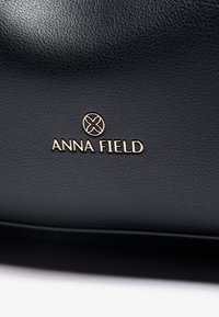 Svart texturerad läderväska med en gyllene "ANNA FIELD"-logotyp, med en slät yta och minimalistisk design.