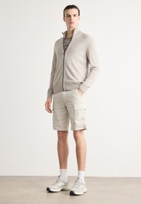 Lys beige zip-up genser med ribbede mansjetter, kombinert med beige cargo shorts med sidelommer og en teksturert stoff. Brukt med grå sneakers.