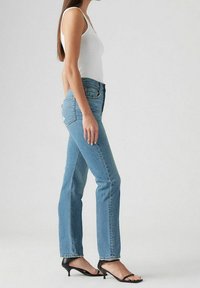 Jean taille haute bleu clair avec une jambe droite, associé à un débardeur blanc et des sandales à talons noires. Design simple, texture en denim classique.