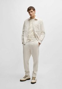 Giacca camicia leggera beige con due tasche, indossata sopra una t-shirt beige chiaro e pantaloni slim coordinati, abbinata a stivali color crema.