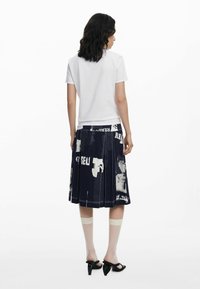 Weißes T-Shirt mit kurzen Ärmeln, kombiniert mit einem marineblauen, plissierten Rock mit einem weißen Grafikdruck. Das Model trägt transparente Socken und schwarze High Heels.