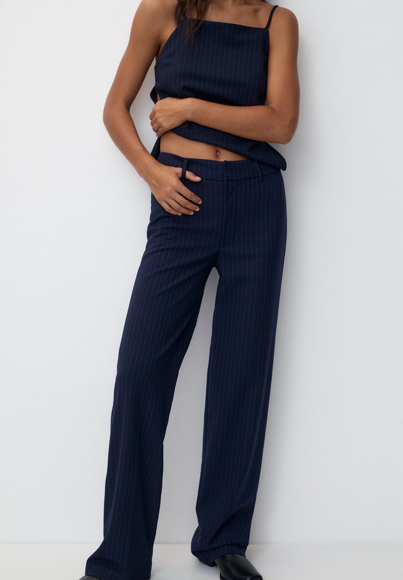 Femme portant un pantalon large à fines rayures bleu marine et un haut sans manches assorti, posant les mains près de la taille devant un fond uni.