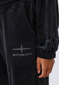 Zwart velours trainingspak met een relaxte pasvorm, voorzien van zilveren strass-accenten op de mouwen en zak. Elastische tailleband en boorden.