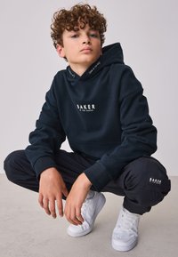 Sort hættetrøje med hvidt "BAKER" logo, løs pasform. Kombineret med sorte bukser og hvide sneakers. Krøllet hår, naturlig hudtone.