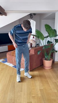 Mėlynos ir žalios juostelės trumpomis rankovėmis marškiniai, šviesiai mėlynos džinsai ir balti sportbačiai. Stovi ant medinių grindų šalia vazono su augalu.