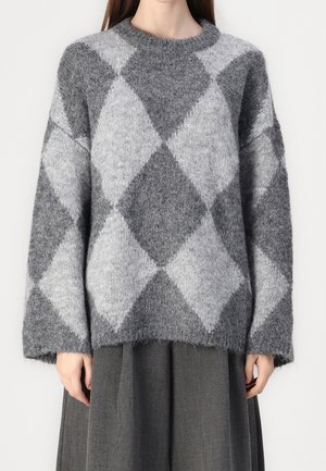 Vrouw in een grijze en lichtgrijze argyle gebreide trui met lange mouwen en een ronde halslijn.