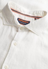 Camicia di lino bianca con colletto, caratterizzata da un'apertura con bottoni e un'etichetta di marca in arancione e blu, con una finitura testurizzata.