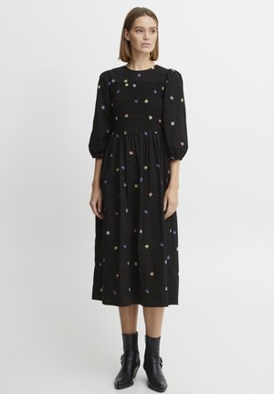 Abito midi nero con corpetto arricciato, maniche a palloncino e ricami floreali multicolori. Indossato con stivaletti neri alla caviglia.