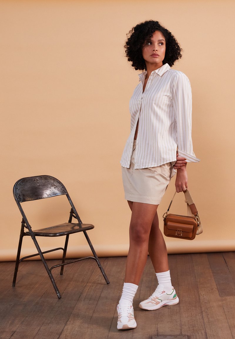 Chemise à rayures blanches, jupe beige, sac à main marron, chaussettes blanches et baskets. Fond avec un sol en bois et une chaise pliante.
