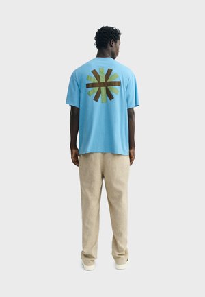 Mann steht mit dem Rücken zur Kamera, trägt ein blaues T-Shirt mit braunem und grünem Sonnenstrahlmuster, beige Hosen und weiße Schuhe vor einem einfarbigen Hintergrund.