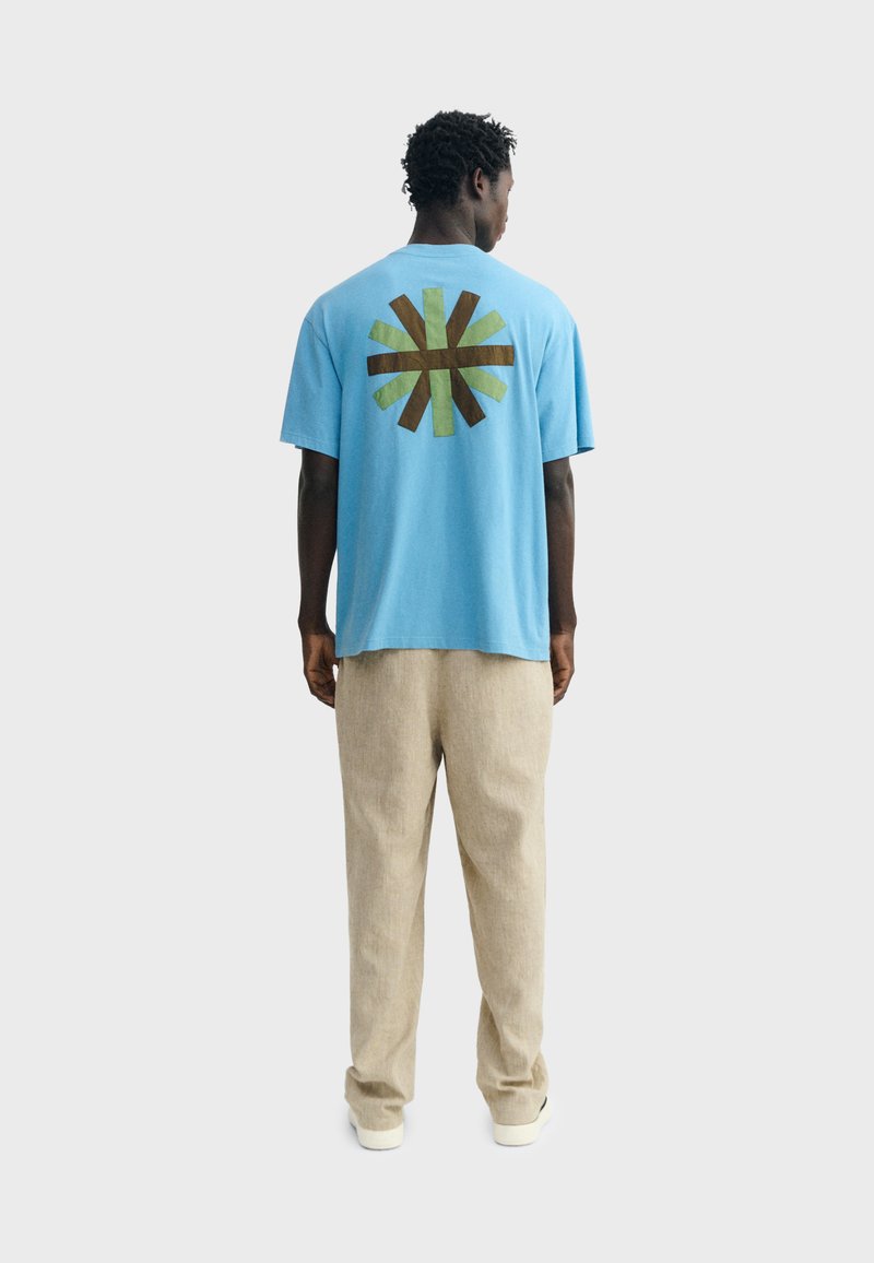 Homme vu de dos, portant un T-shirt bleu avec un motif d'explosion de soleil marron et vert, un pantalon beige et des chaussures blanches sur un fond uni.