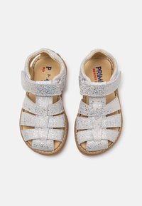 Glitzernde silberne Sandalen mit überkreuzten Riemen, verstellbarem Klettverschluss und einer sandfarbenen Gummisohle. Strukturierter Oberstoff mit schimmernden Akzenten.