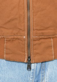 Brun zip-up jakke med en tekstureret overflade, der har en metallynlås og hvid stikning. Sammen med blå denim nederst.