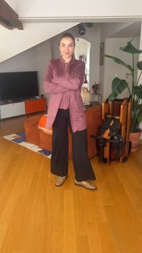 Femme en veste mauve et pantalon noir debout dans un salon avec un canapé orange, une grande plante et un rameur en bois à côté d’elle.