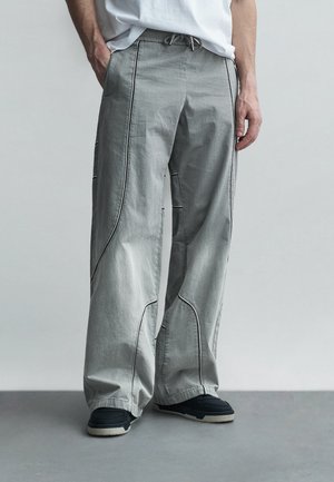 Personne portant un pantalon ample gris clair avec des accents de couture noirs, associé à des baskets noires et blanches, debout sur un sol gris.