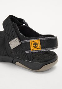 Μαύρη σαγιονάρα με ανθεκτικό ύφασμα στην επάνω πλευρά, γκρίζες λεπτομέρειες, ρυθμιζόμενη λωρίδα, υφή στο πέλμα και έντονη κίτρινη ετικέτα με λογότυπο.