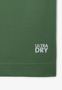 Tela de color verde oscuro con textura y un patrón de malla; presenta un logo blanco que dice "ULTRA DRY" cerca de la esquina inferior derecha.