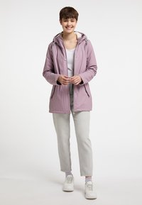 Giacca impermeabile di un leggero viola con cappuccio, chiusura a snap e zip, due tasche frontali, indossata sopra una maglietta bianca e pantaloni grigio chiaro.