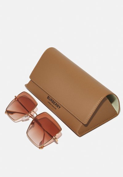 Kwadratowe okulary przeciwsłoneczne z brązowymi gradientowymi soczewkami i złotymi oprawkami obok brązowego skórzanego etui na okulary przeciwsłoneczne Burberry.