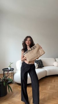 Sac de cumpărături din hârtie maro cu logo negru, ținut de o persoană îmbrăcată în ținută neagră cu dungi verticale, amplasat într-un interior modern.