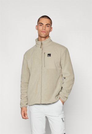 Veste en polaire beige avec un col haut, poche poitrine zippée et patch logo. Manches longues et ourlet droit, texture douce.