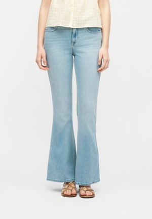 Žena v svetlomodrých rozšírených džínsoch, béžovom tielku bez rukávov a hnedých sandáloch s remienkami, stojaca pred jednoduchým svetlým pozadím.