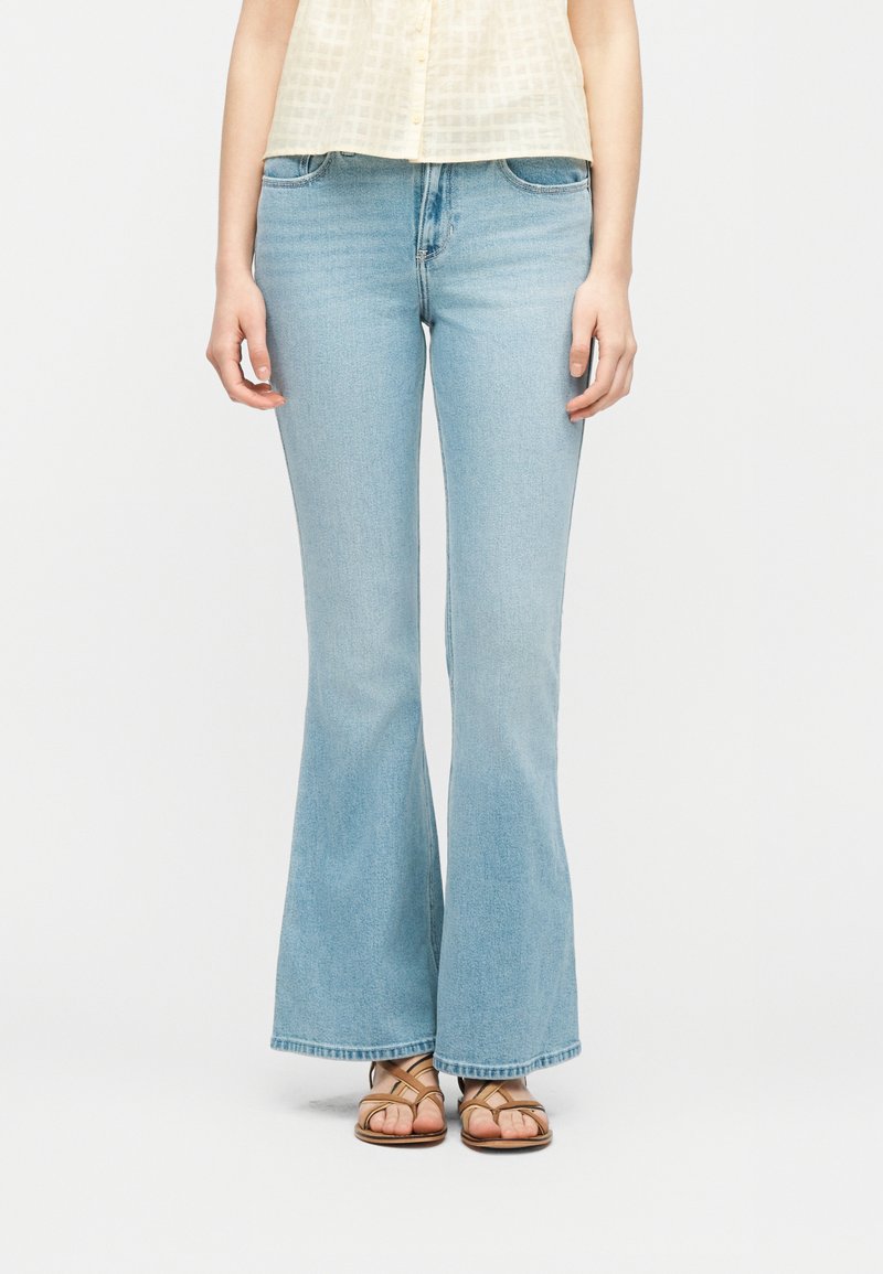 Γυναίκα που φορά ανοιχτό μπλε φαρδιά τζιν, μπεζ αμάνικο τοπ και καφέ σανδάλια με λουράκια, όρθια μπροστά σε απλό ανοιχτό φόντο.