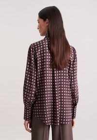 Braun gemustertes Blusenshirt mit Puffärmeln, das cremefarbene quadratische Motive aufweist. Das Material hat eine glatte Textur und einen lockeren Schnitt. Rückansicht angezeigt.