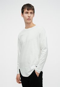 Ung mand med kort mørkt hår, iført en ensfarvet hvid langærmet skjorte og sorte bukser, står med den ene hånd i lommen.