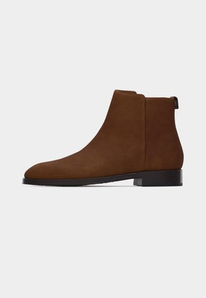 Smeđa gležnjača od brušene kože s niskom petom, zaobljenim vrhom i trakom za povlačenje na stražnjoj strani, prikazana iz profila na bijeloj pozadini.