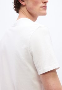 Άνδρας που φοράει απλό λευκό κοντομάνικο T-shirt, εμφανιζόμενος από την πίσω και πλάγια όψη πάνω σε απλό λευκό φόντο.