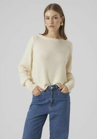 Krämfärgad stickad tröja med diamantmönster, bred halsringning och ribbade manschetter, kombinerad med högmidjade blå jeans.