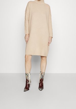 Femme portant une robe pull en maille beige avec des manches torsadées et des bottines à talons hauts en marron à motif peau de serpent.