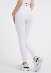 Žena v texturovaných bílých legínách a odpovídajícím crop topu, pózující s jednou nohou mírně zvednutou, v bílých a černých teniskách.