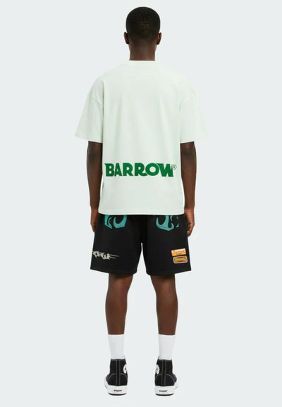 Mann, der mit dem Rücken zur Kamera steht, trägt ein weißes Barrow-T-Shirt und schwarze Shorts mit bunten Aufnähern, weiße Socken und schwarze High-Top-Sneaker.