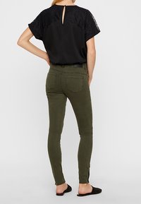 Top nero con dettagli in pizzo e apertura a chiave sul retro, abbinato a jeans skinny verde oliva. Calzature scure completano l'outfit.