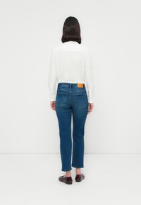 Kvinne med skulderlangt mørkt hår iført en hvit langermet skjorte, blå jeans og mørkebrune flats, står vendt bort fra kameraet mot en hvit bakgrunn.