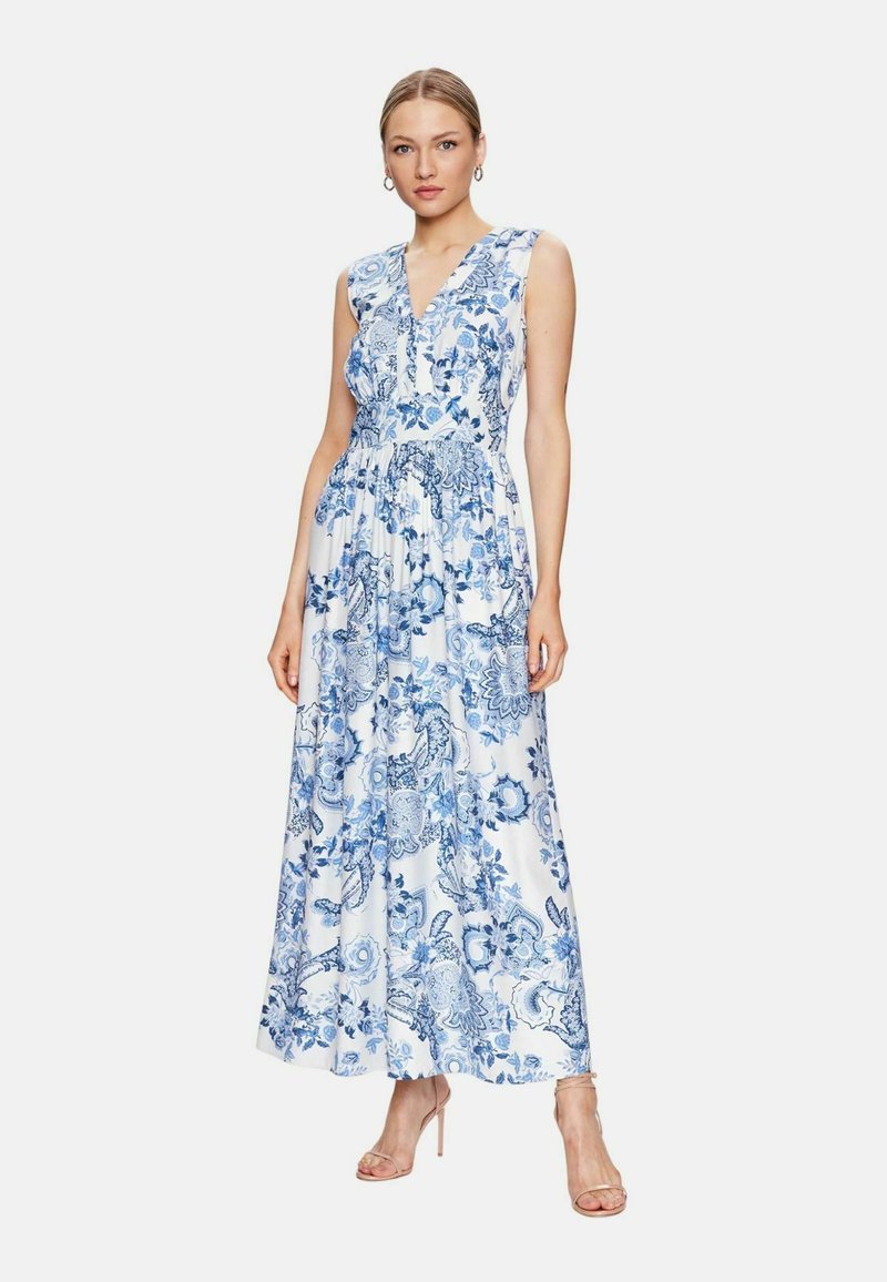 Abito maxi, tessuto bianco con motivi floreali e paisley blu, design senza maniche, scollatura a V, vita aderente, gonna fluida, indossato con sandali con cinturino.