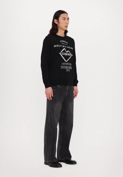 Sweat-shirt noir avec texte blanc, associé à un jean large gris foncé avec des coutures latérales. Le modèle se tient devant un fond uni.